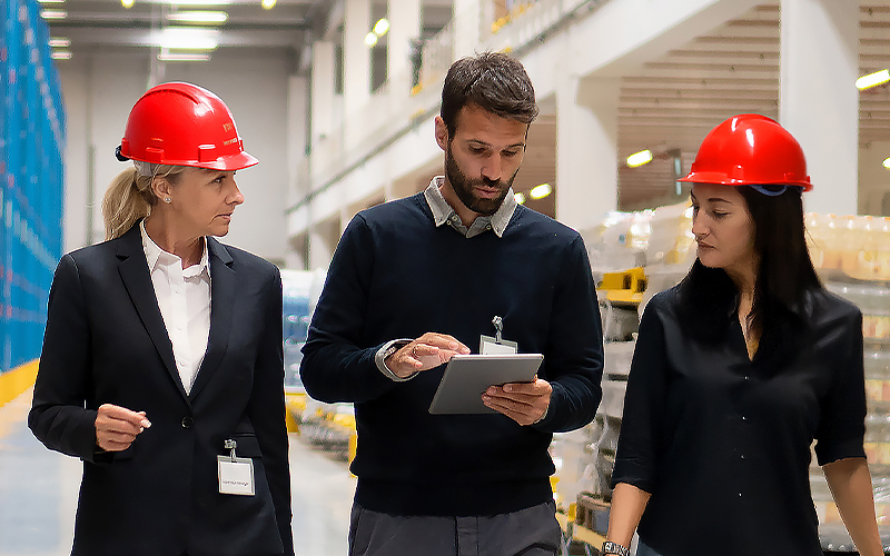 Three people wearing red safety helmets and business attire stand in a warehouse while one person uses a tablet and the others watch attentively.