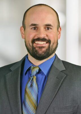A man with a beard, wearing a gray suit, blue shirt, and striped tie, smiles at the camera against a blurred light background.