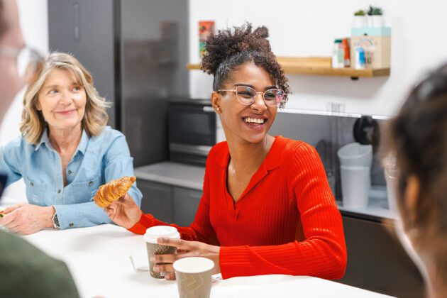 A woman in a red sweater smiles while holding a coffee cup and croissant, sitting at a table with others in a modern kitchen or break room.