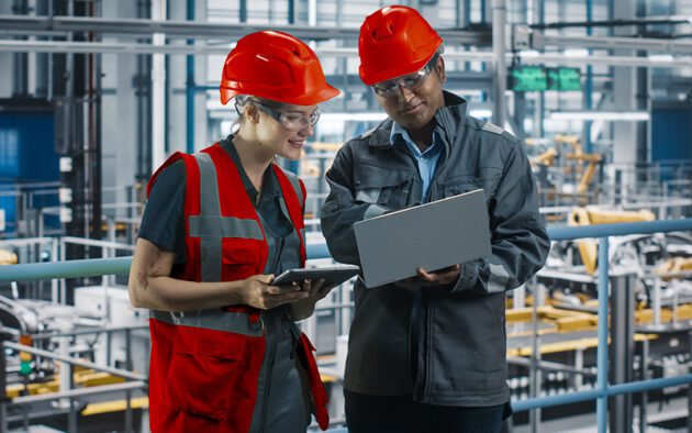 Two industrial workers wearing safety helmets and vests review information on a tablet and laptop in a factory setting.