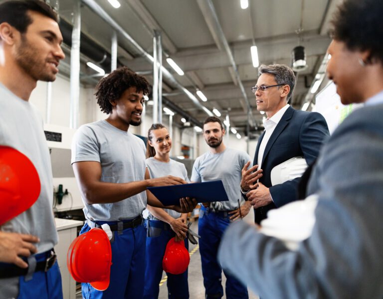 A group of workers in blue uniforms listen to a person in a suit holding a helmet, while another worker holds a clipboard in a factory setting.
