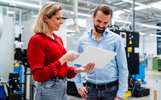 Two people in business casual attire review information on a tablet in a modern industrial or factory setting with machinery in the background.