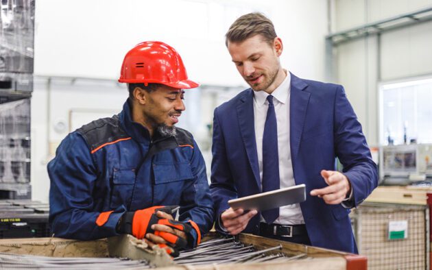 A factory worker in safety gear and a man in a suit discuss something on a tablet beside a box of metal rods in an industrial setting.