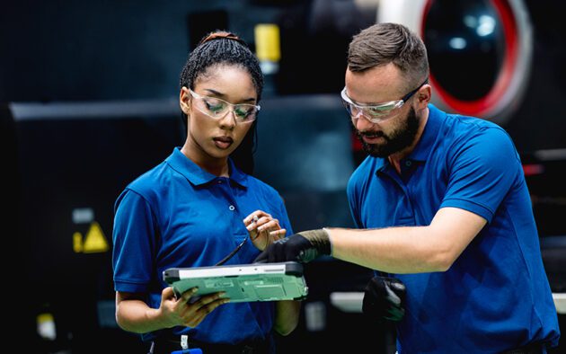 Two factory workers in blue uniforms and safety glasses review information on a tablet together in an industrial setting.