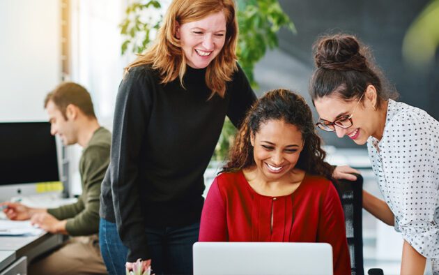 Three women smiling and looking at a laptop together in an office, while a man works at a desk in the background.