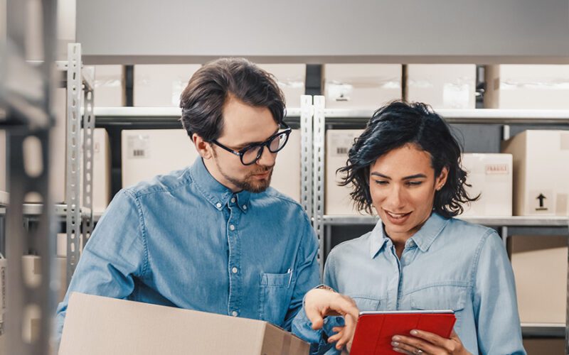 Two people in a warehouse setting look at a tablet together. One holds a cardboard box while the other points at the screen. Shelves with packages are visible in the background.