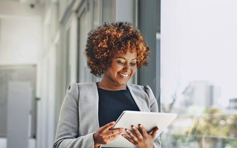 A woman in business attire stands by a window, smiling while using a digital tablet.
