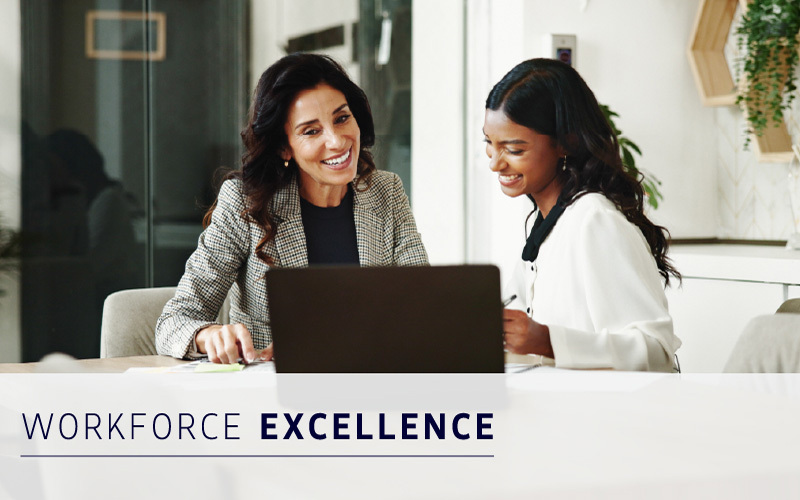 Two professionally dressed women sit at a table, smiling and working together on a laptop. Text overlay reads: "Workforce Excellence.