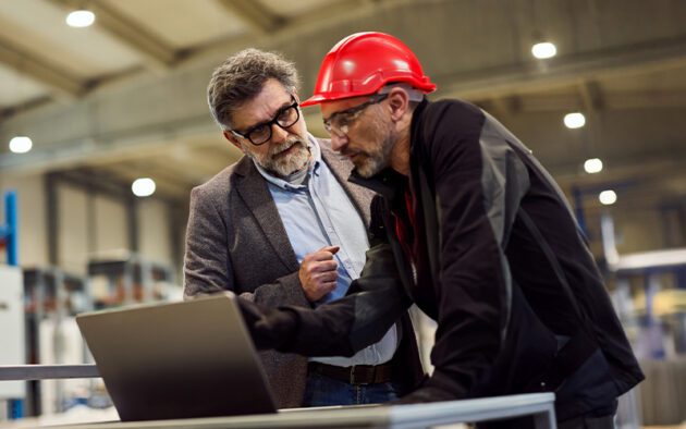 Two men discussing work at a factory; one wears a red hard hat and jacket, the other wears glasses and a blazer. They are looking at a laptop on a metal surface.