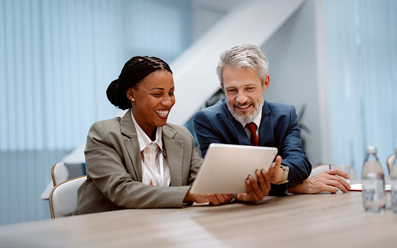 Two business professionals sit at a table, smiling and looking at a tablet together in a modern office setting.