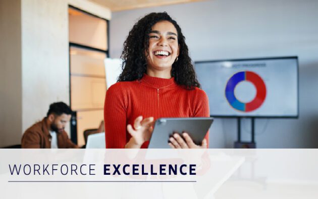 A woman in a red sweater smiles while holding a tablet in an office; a man works at a desk and a screen displays a pie chart in the background. Text reads "Workforce Excellence.