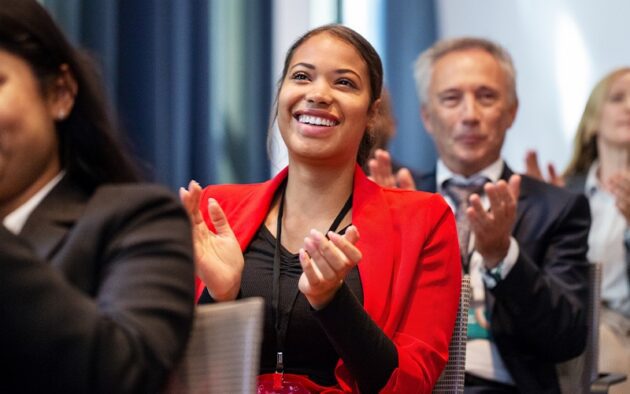 A woman in a red blazer is smiling and clapping in an audience, with other people around her also applauding.