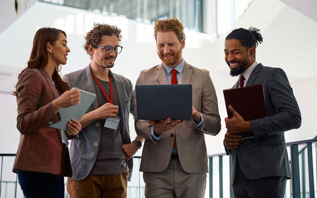 Four professionals, two men and two women, stand together indoors, smiling and looking at a laptop held by one man; they appear to be in a business or office setting.