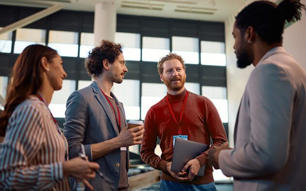 Four people wearing conference badges stand indoors having a conversation; one holds a laptop, another has a coffee cup, and they appear engaged and attentive.