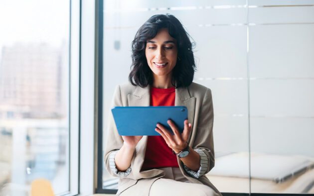 A woman in business attire sits by a window, holding and looking at a tablet with a slight smile.