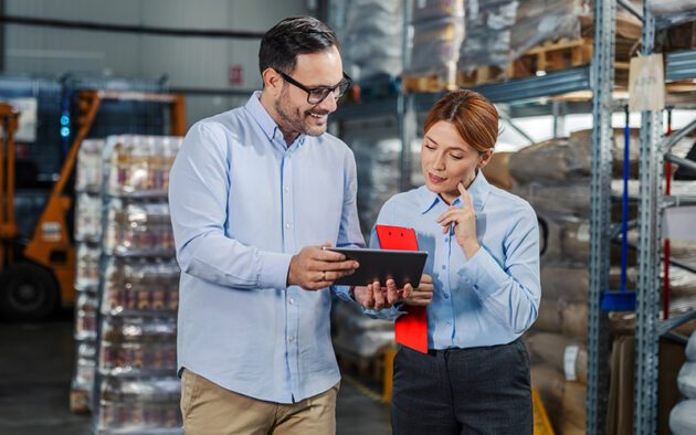 Two people in business attire review information on a tablet while standing in a warehouse with shelves stocked with goods in the background.