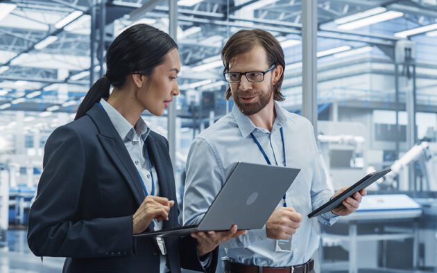 Two professionals stand in a modern industrial setting, discussing information displayed on a laptop and a tablet.
