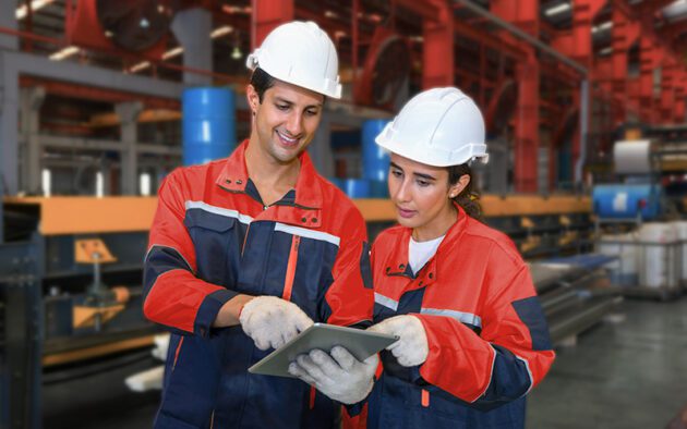 Two workers wearing safety helmets and gloves look at a tablet in a factory setting, surrounded by industrial equipment and red structural beams.