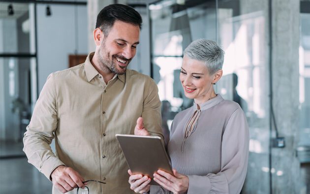 A man and a woman standing together in an office, smiling while looking at a tablet held by the woman.
