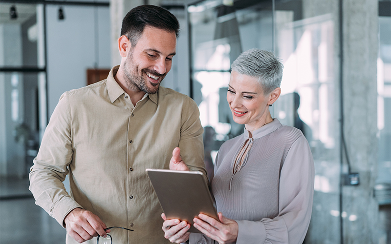 A man and a woman standing together in an office, smiling while looking at a tablet held by the woman.