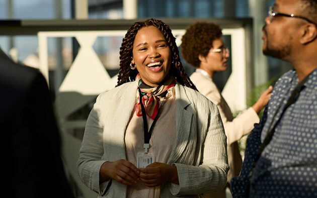 A woman wearing a lanyard and a scarf smiles while standing indoors among other people in business attire.