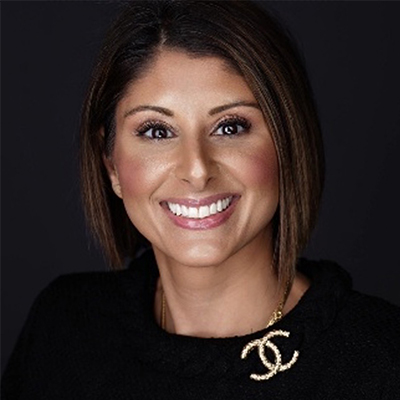 Woman with straight, shoulder-length brown hair, wearing a black top and a gold Chanel necklace, smiling against a dark background.