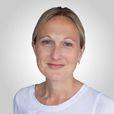 A woman with light hair pulled back, wearing a white shirt, smiling softly in front of a plain, light gray background.
