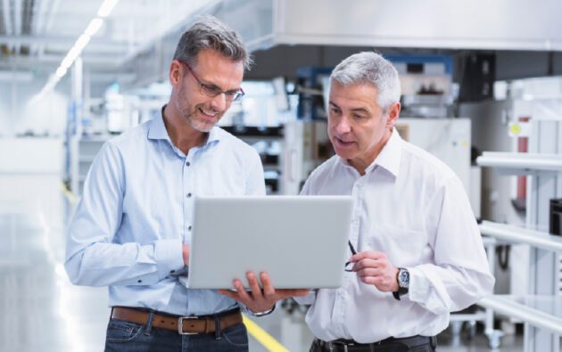 Two men in business attire standing in a modern industrial setting, looking at a laptop screen and discussing its contents.