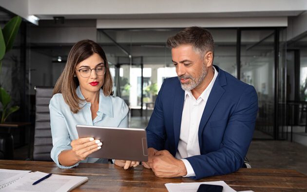 Two business professionals sitting at a desk, reviewing information on a tablet in a modern office setting.