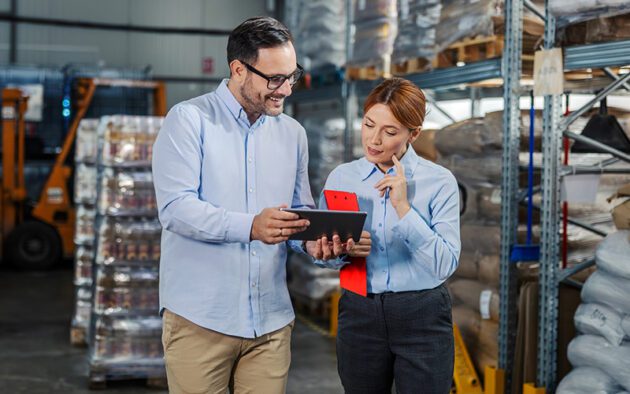 Two people in business attire stand in a warehouse, reviewing information on a tablet and clipboard, surrounded by shelves with goods and packaged products.