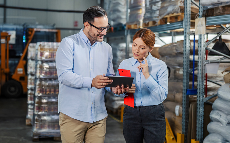 Two people in business attire stand in a warehouse, reviewing information on a tablet and clipboard, surrounded by shelves with goods and packaged products.