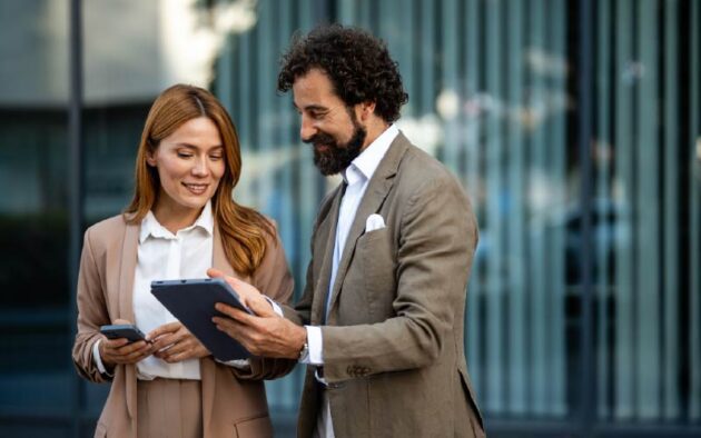 Two business professionals in suits standing outdoors, one holding a phone and the other showing something on a tablet, both appearing engaged in conversation.