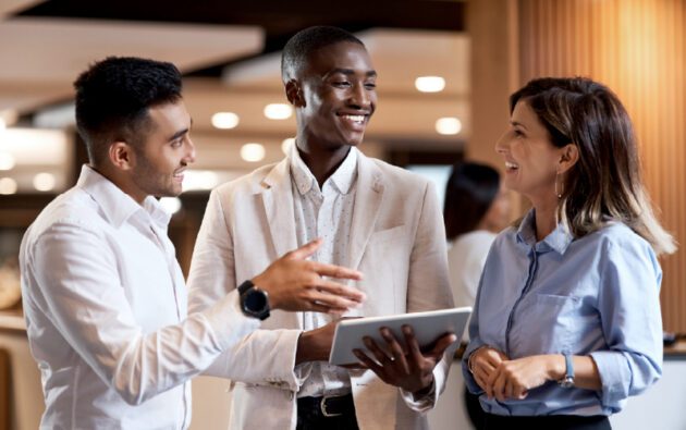 Three people in business attire stand indoors, smiling and talking. One holds a tablet while gesturing, and the others listen attentively.