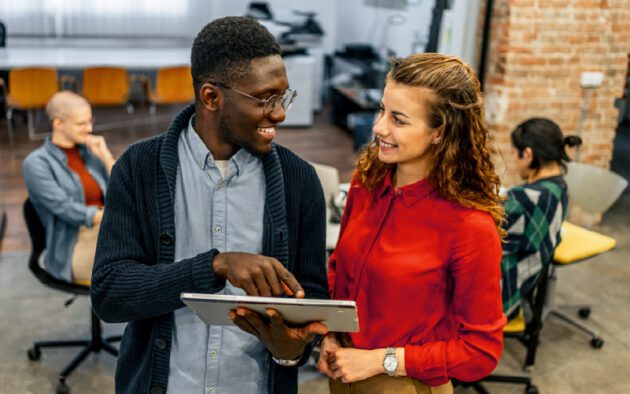 Two colleagues stand and discuss something on a tablet while smiling; other people work in the background in a modern office space.