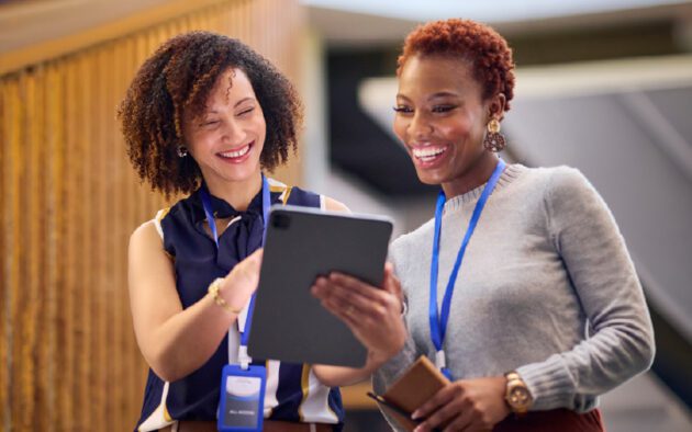 Two women wearing lanyards stand indoors, smiling and looking at a tablet device together.