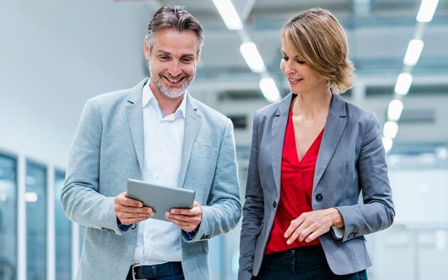 Two business professionals, a man and a woman, stand indoors. The man holds a tablet and smiles while the woman looks at the tablet. Both are dressed in business attire.