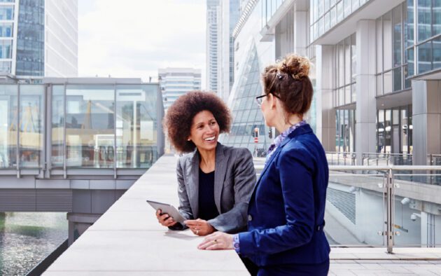Two women in business attire have a conversation outdoors in a modern urban setting; one holds a tablet while they both smile.