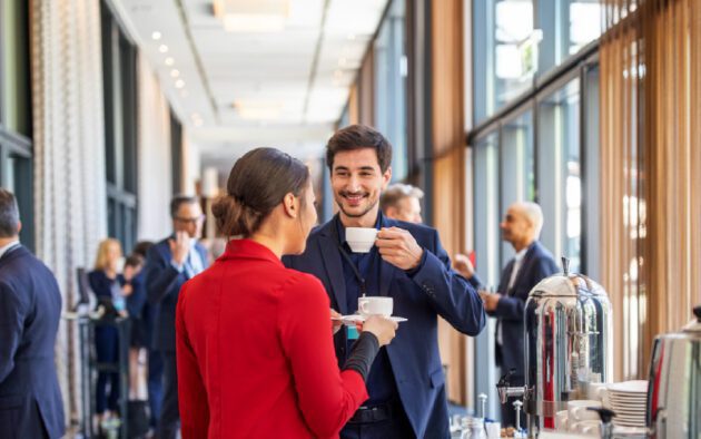 Two people in business attire hold coffee cups and talk at a networking event in a bright, modern conference space with other attendees in the background.