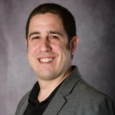 A man with short dark hair smiles at the camera, wearing a gray blazer over a black shirt, with a neutral gray background.