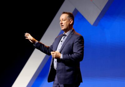 A man in a suit speaks onstage, gesturing with one hand, in front of a blue geometric background.