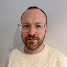 A man with short reddish-brown hair, a beard, and glasses wearing a cream-colored sweater stands in front of a plain light gray wall—reflecting the diversity seen in today's global workforce.