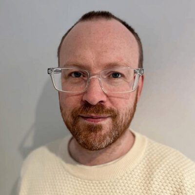 A person with short reddish-brown hair, a trimmed beard, and clear rectangular glasses is wearing a cream-colored textured sweater and standing against a plain light background.