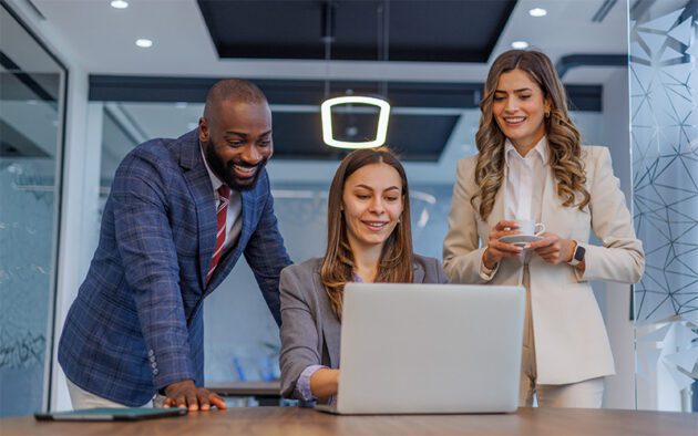 Three professionals in business attire look at a laptop screen together in a modern office, with one person holding a coffee cup and another pointing at the laptop.