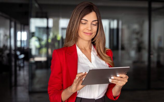 Woman in a red blazer stands indoors, engaging with her tablet device—connected to the global workforce from anywhere.