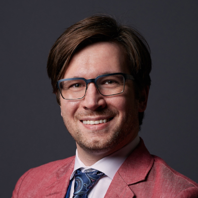 A man with brown hair and glasses smiles at the camera. Dressed in a pink blazer, white shirt, and blue patterned tie, he exudes confidence—perfect for a workforce management professional—against a dark background.