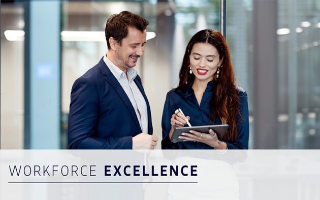 Two professionally dressed people stand indoors, one holding a tablet and stylus. Both are smiling, reflecting effective workforce management. Text overlay reads “Workforce Excellence.”.