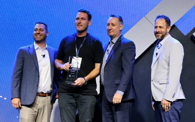 Four men in business attire stand together on stage, one holding a workforce management award, all facing the camera and smiling, with a blue backdrop behind them.
