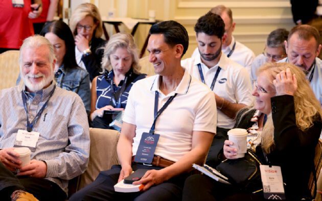 A group of people sit in rows at a conference, some smiling and holding coffee cups, all wearing name badges and conference lanyards at a workforce management event.
