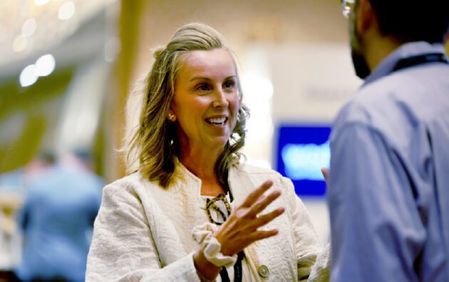 A woman in a light-colored jacket gestures while discussing workforce management with a man wearing glasses and a blue shirt indoors.