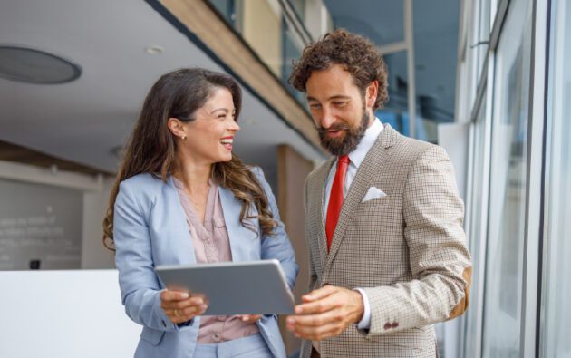 Two business professionals standing indoors, looking at a tablet and smiling, with large windows in the background.